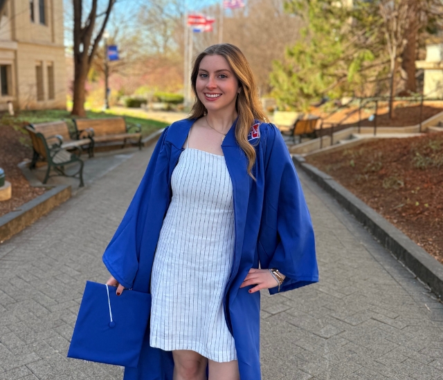Photo of student in AU blue graduation gown holding grad cap with campus in background.