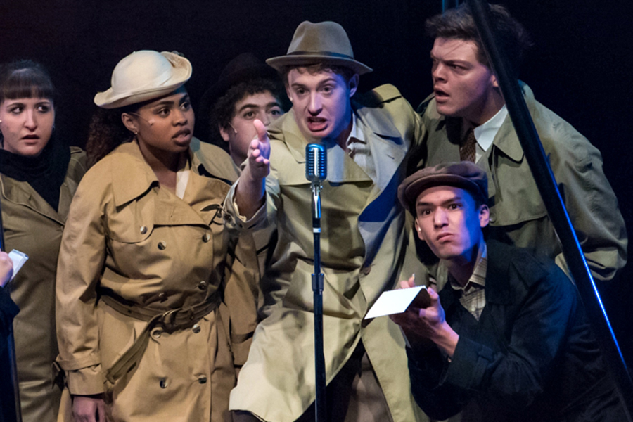 A group of people in trench coats cluster around a microphone