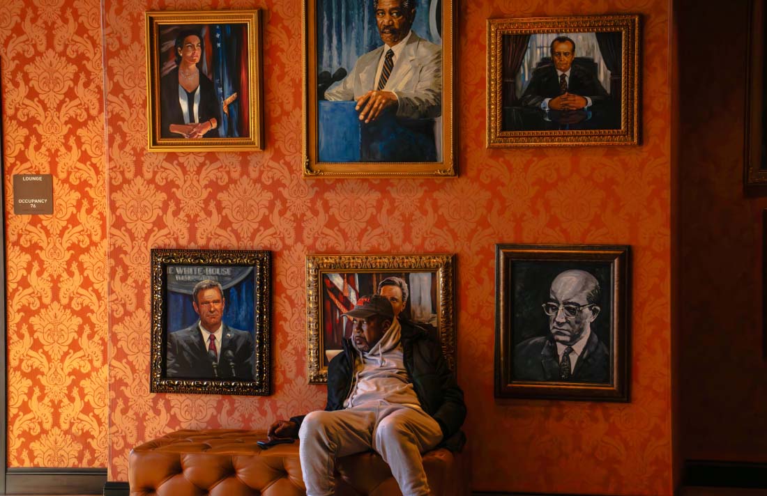 Photo: Mads Ernst (CAS/SOC ‘27), American Gaze, 2025. A man in casual clothes sitting on tufted bench under painted portraits in gold frames on damask wallpaper wall.