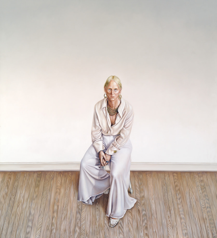 Portrait of a person in flowy white clothing and jewelry in front of a large, blank white wall