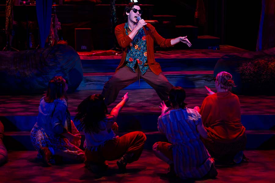 An actor in sunglasses sings into a microphone; other kneeling actors watch him.