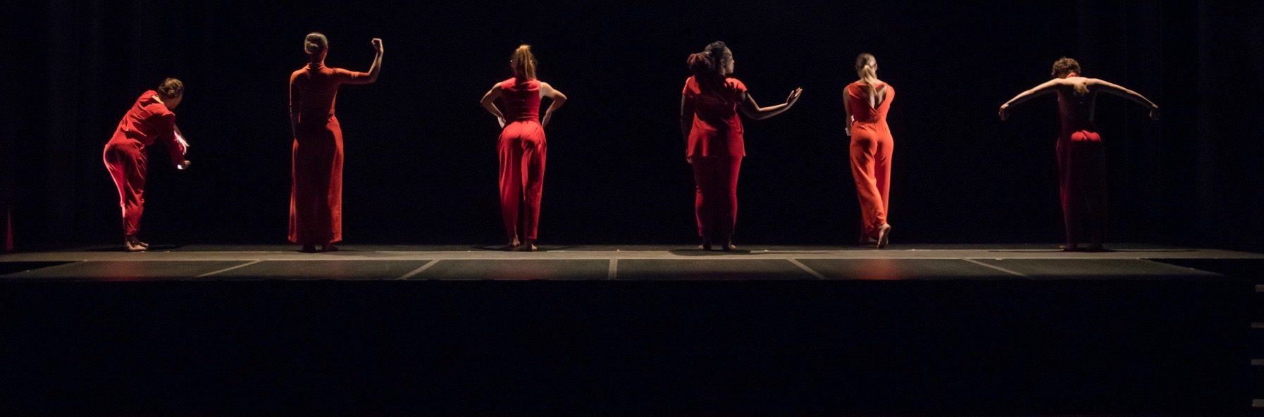 Dancers stand in a line, dressed in red, gesturing towards upstage