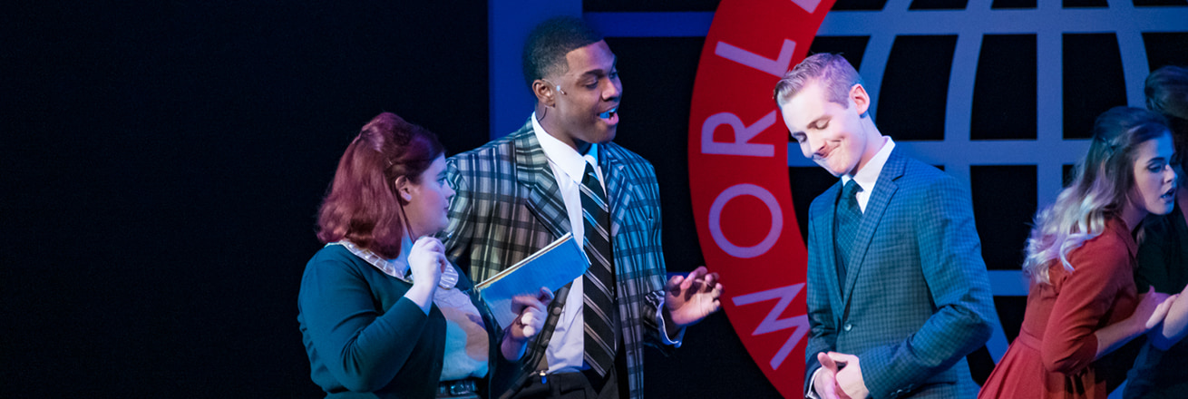 Onstage, people dressed as 1960s office workers have a casualchat