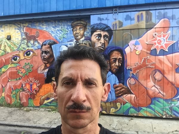 Jose Medina in front of mural depicting people struggling in the grip external forces (symbolized by a hand larger than the people surrounding them).
