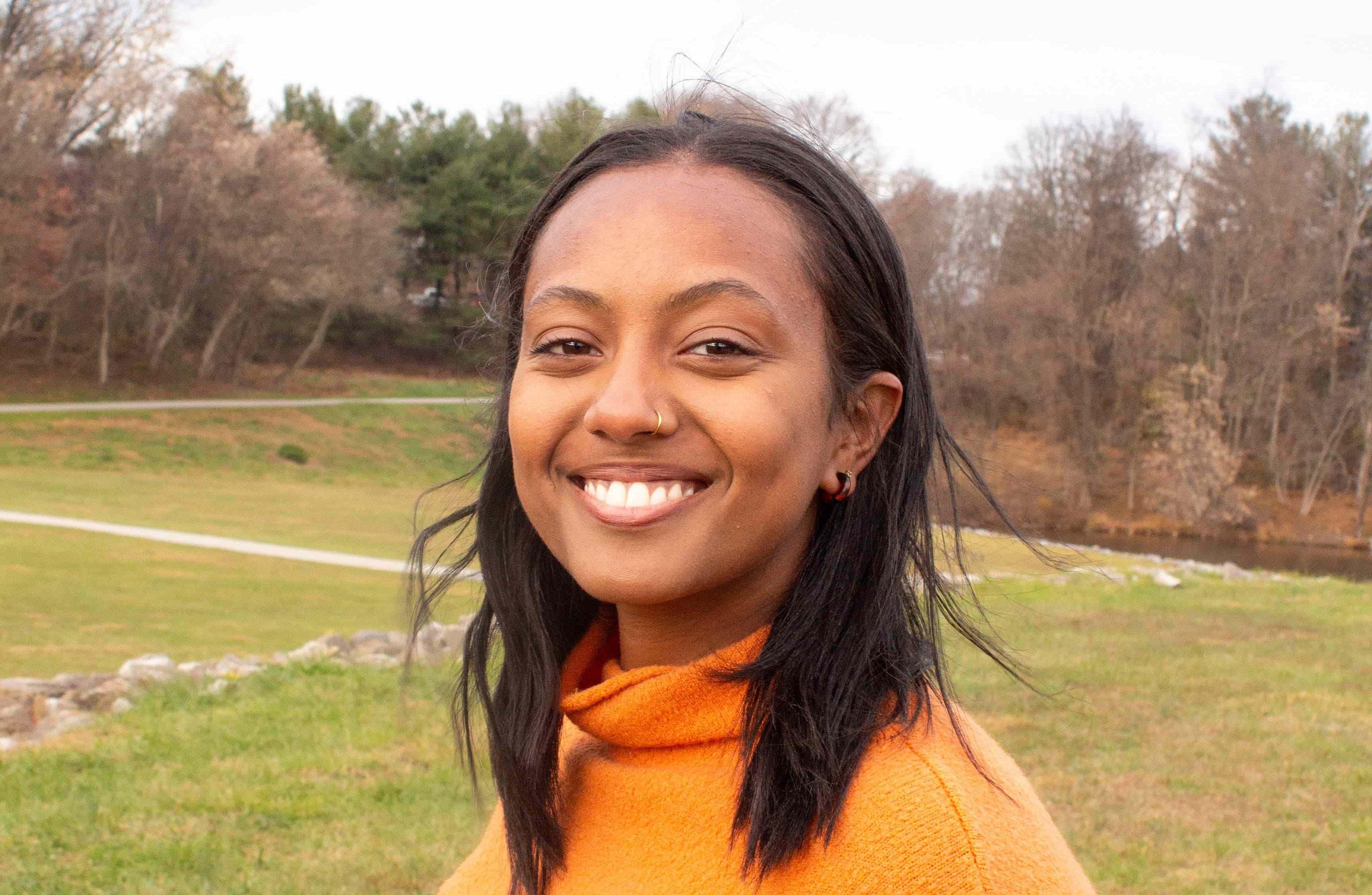 A smiling woman with medium-length black hair wearing an orange sweater, standing outdoors on a grassy field with trees in the background on a cloudy day.