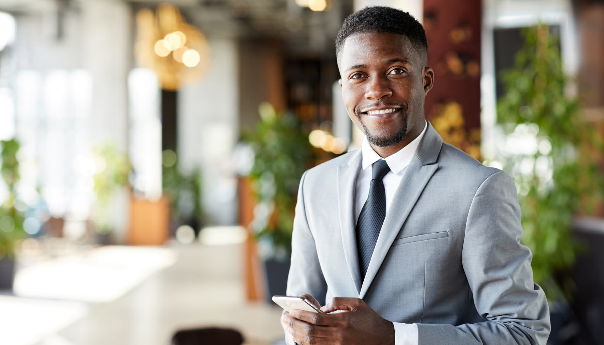 confident young man in a light gray suit and black tie smiles while holding a smartphone in a bright, modern office