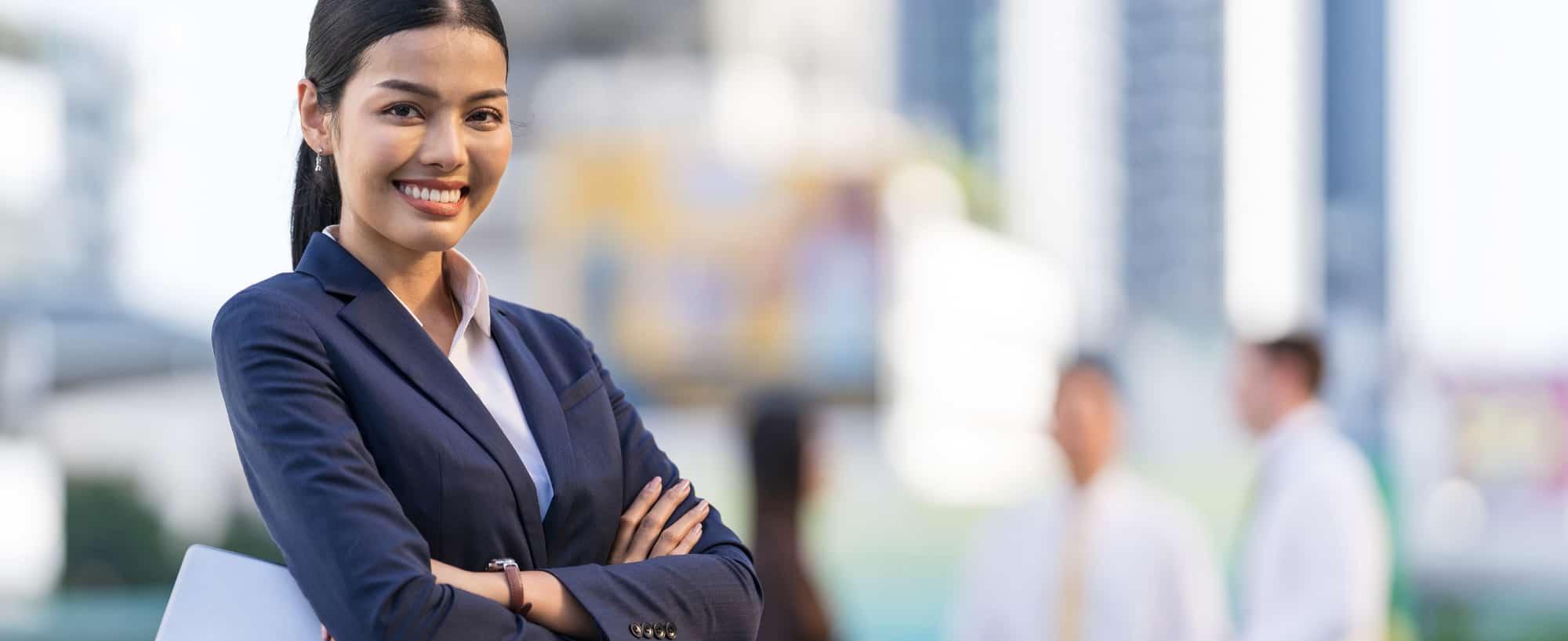 A confident young businesswoman stands outdoors with arms crossed, smiling at the camera. She is dressed in a navy blue suit and holding a tablet.