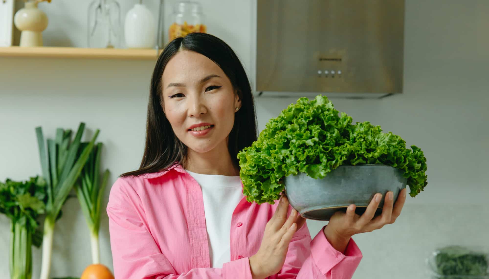 A woman in a pink shirt standing in a kitchen, holding a bowl full of fresh green lettuce and smiling slightly at the camera. Fresh vegetables are visible in the background.