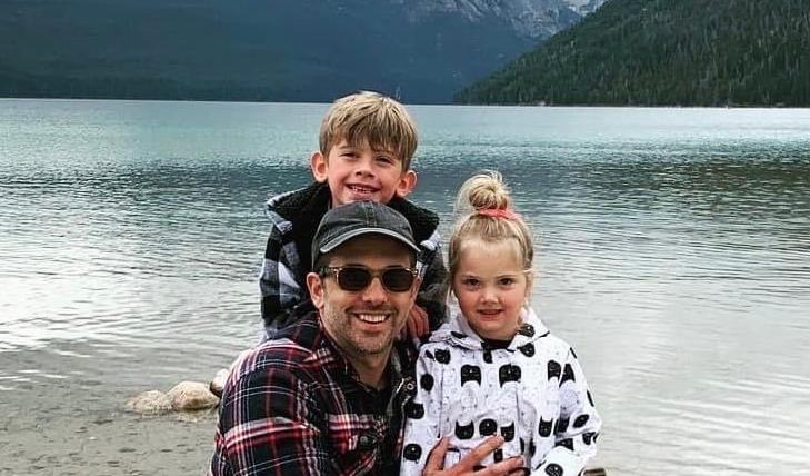 A smiling man crouches by a lake with two young children, one boy and one girl, standing close beside him. They are all dressed warmly in jackets, and the background features a serene mountain lake with pine-covered hills and rugged peaks under a cloudy