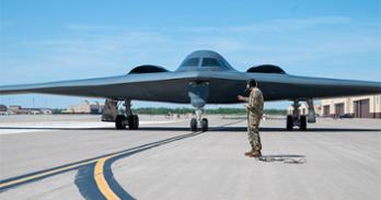 an army man dressed in camo univofrm with headset stands in front of a B2 stealth bomber