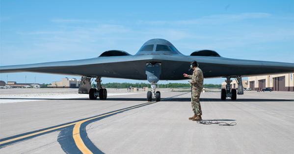 an army man dressed in camo univofrm with headset stands in front of a B2 stealth bomber