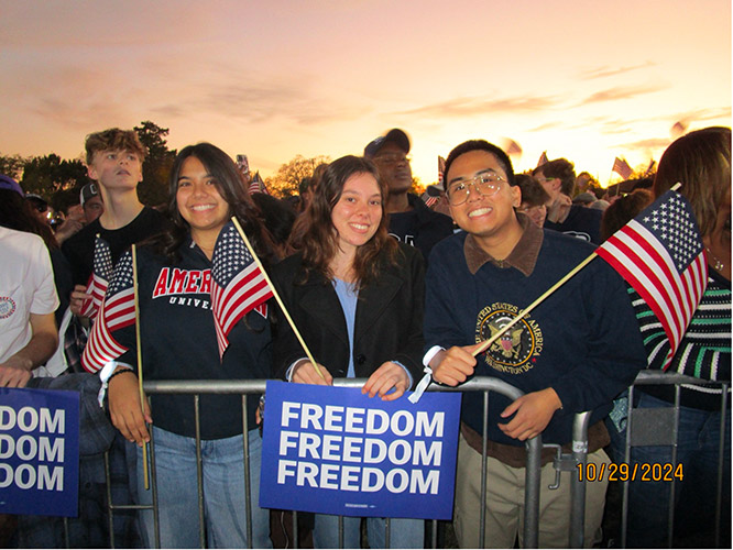 3 students pose at the gate with American flags and a Freedom sign at the Kamala Harris for President rally held on the National Mall