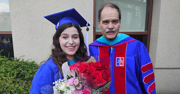Tom and Ava outside SIS. Ava is dressed in graduation regalia and holding flowers. Tom is also in academic robes.
