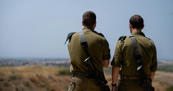Two military officials in green uniforms have their back turned to the camera as they oberve a view