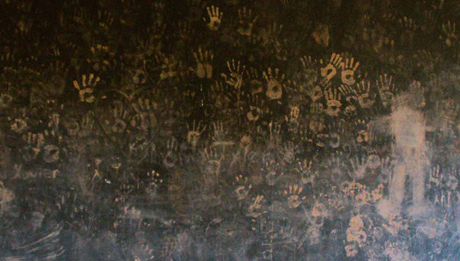 A dark wall with reddish brown handprints covering it. The handprints grow more smudged further down the wall. To the right stands an abstract figure painted with the same reddish color.