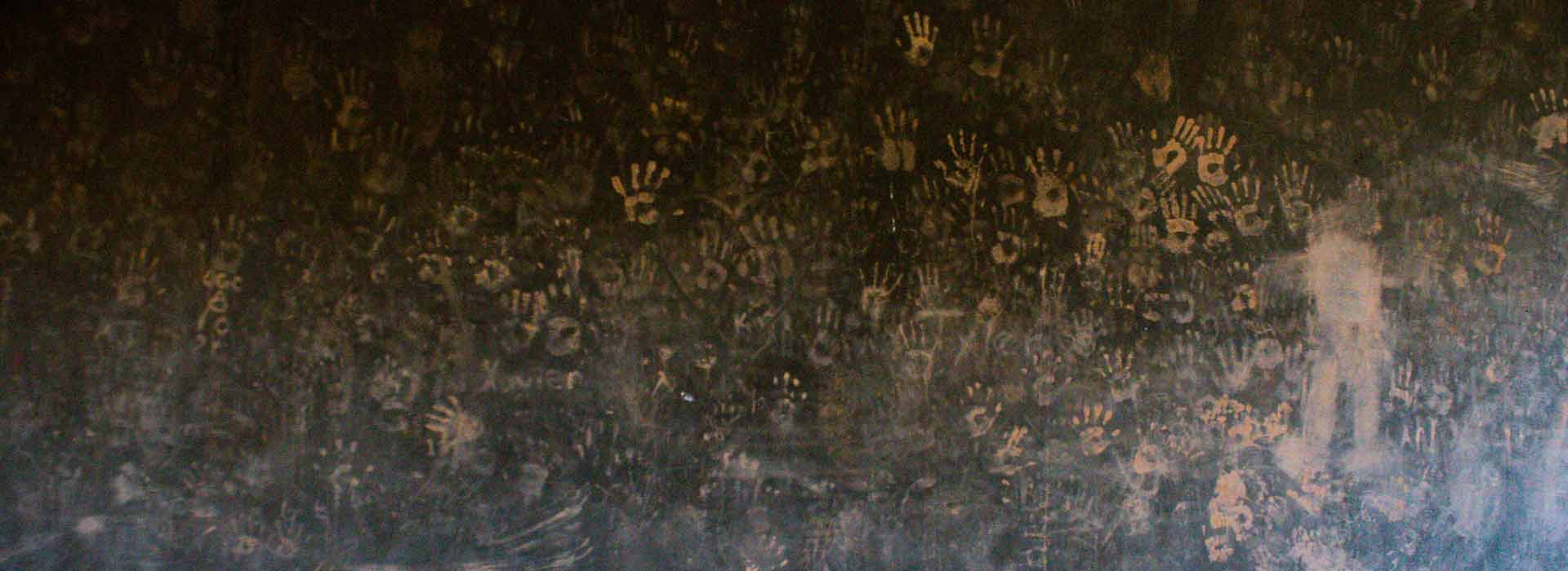 A dark wall with reddish brown handprints covering it. The handprints grow more smudged further down the wall. To the right stands an abstract figure painted with the same reddish color.