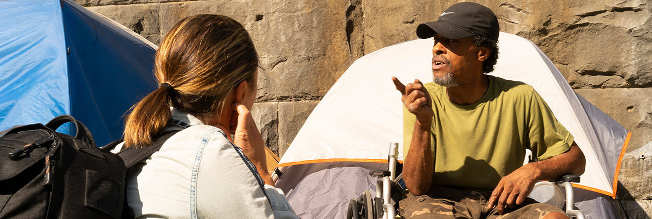 A woman interviewing a homeless man in front of a tent