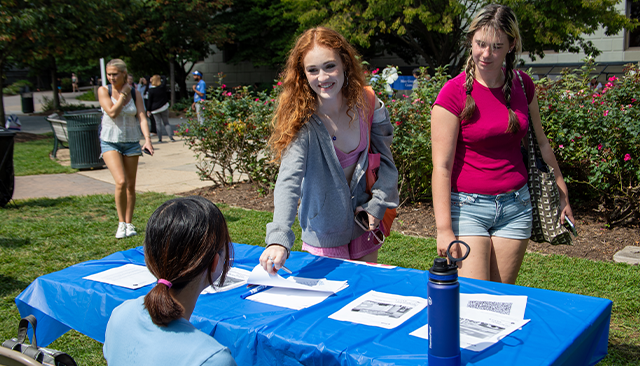 One student reaches for a piece of paper at a table during the Involvement fair. Another student looks at the table while a third student sits at the table with their back to the camera.