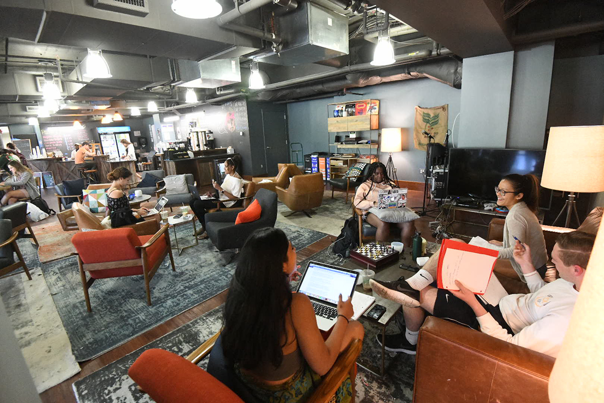 Students sit in the Bridge Cafe, working on their laptops in several orange chairs.