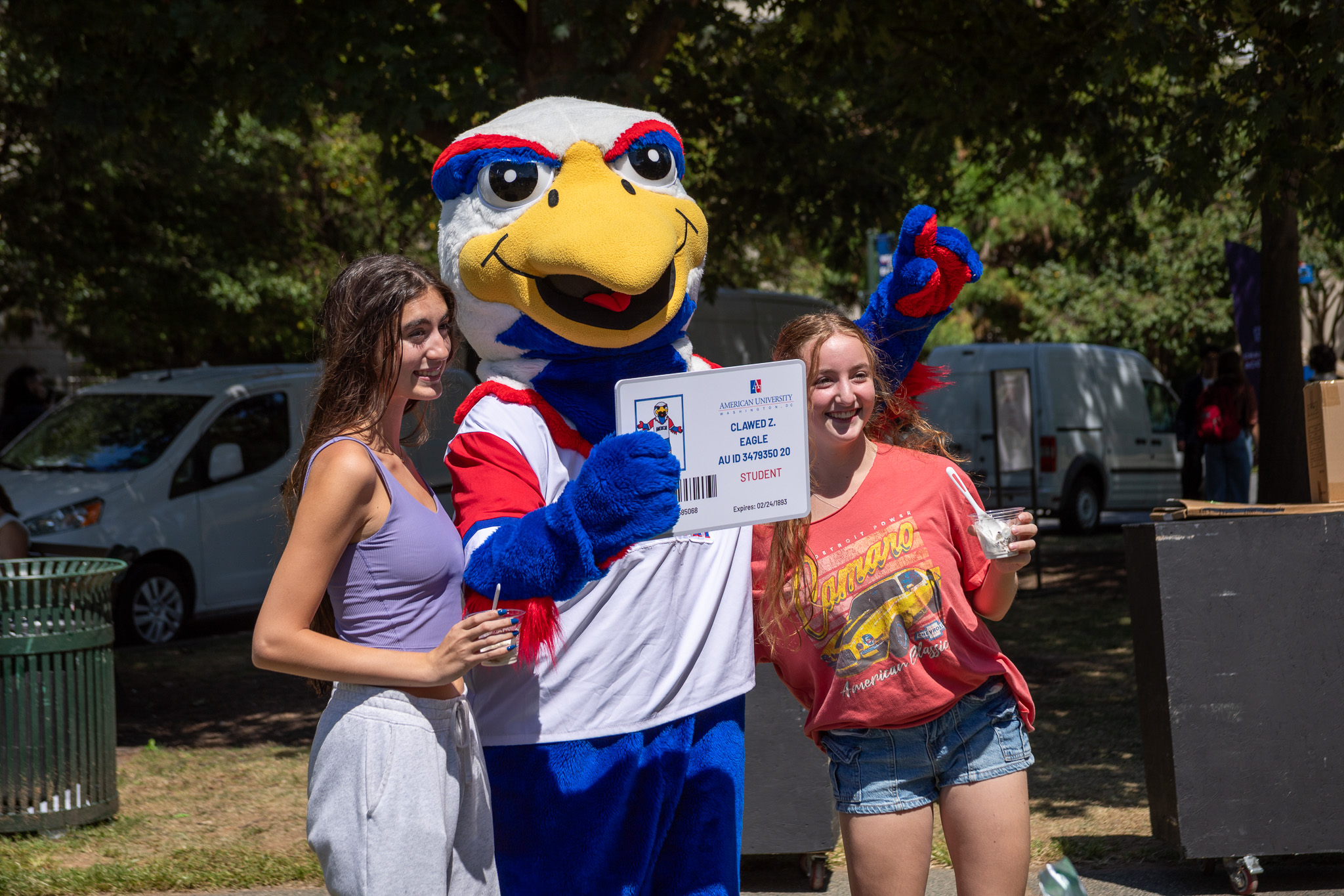 Two students pose with Clawed, AU's eagle mascot, on the Quad. Clawed holds a sign that looks like a student ID, while the two students hold cups with ice cream.